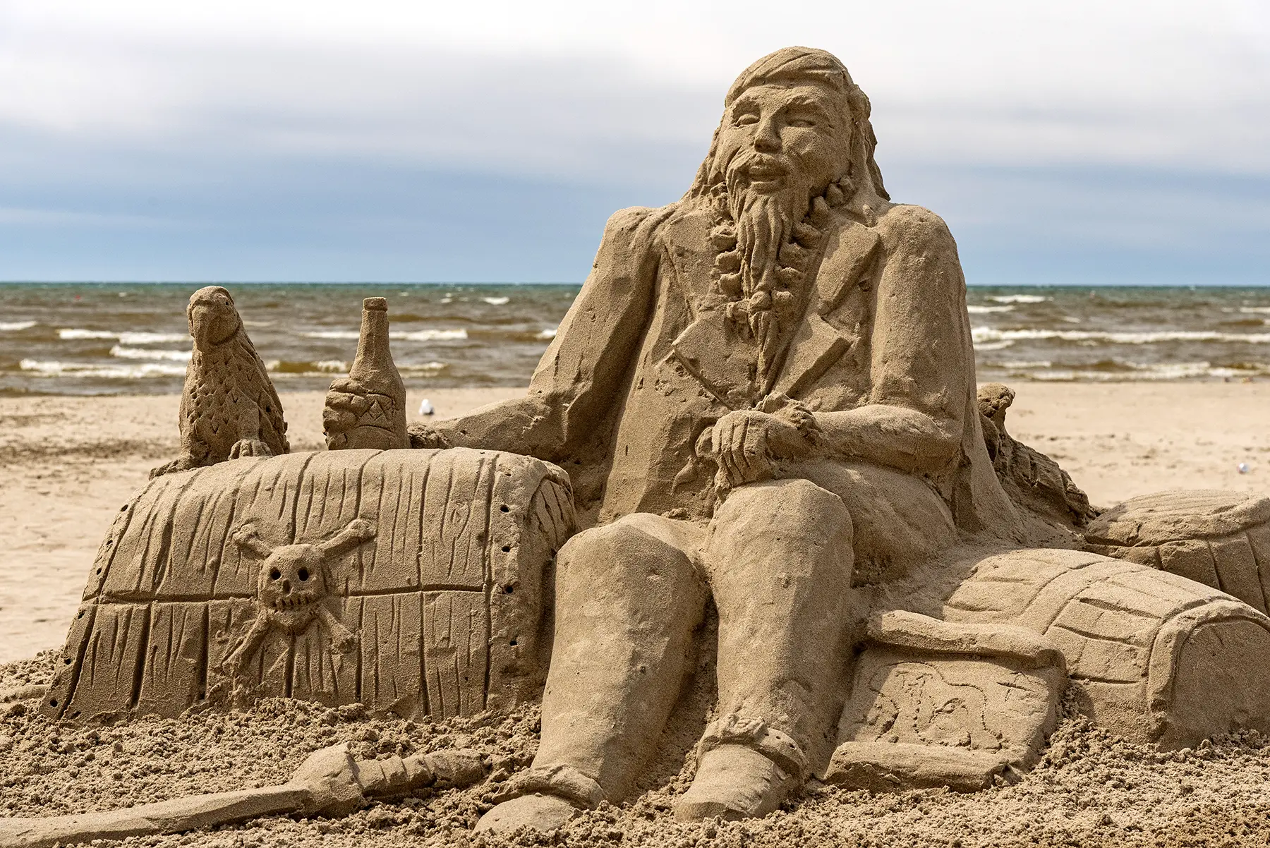 Life-size sand sculpture of a bearded man seated on a beach, with a carved ship wheel and bottle nearby in the sand.