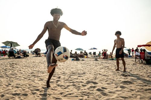 Two shirtless young men playing beach soccer on a crowded sunny beach; one kicks the ball toward the camera.