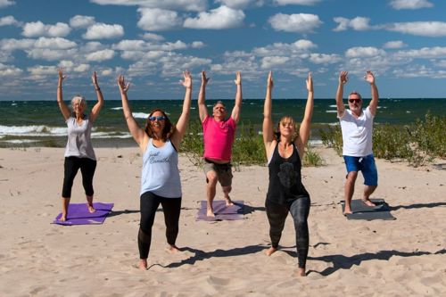 Five adults stand on a sunny beach with arms raised in a yoga pose, ocean and clouds behind them.