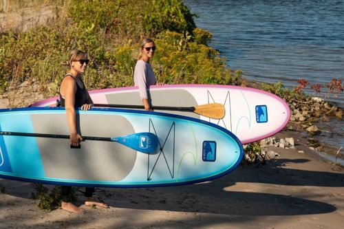 Two women stand on a sandy beach by the water, carrying colorful stand-up paddleboards (blue foreground, pink in back).