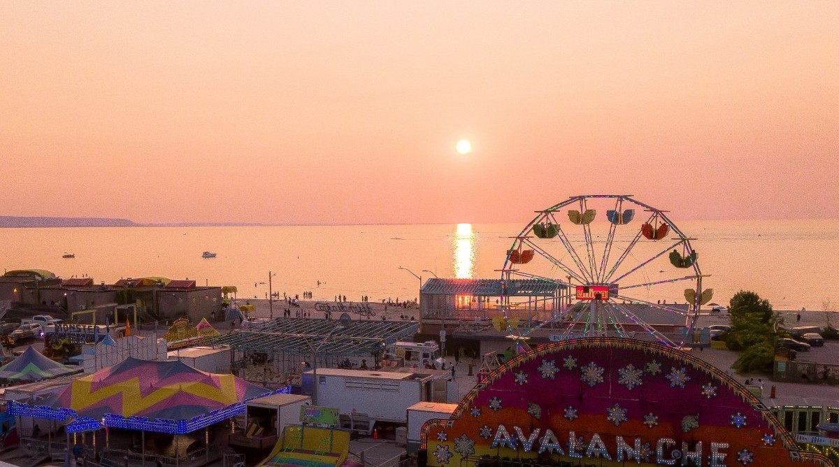 Sunset over a seaside carnival with a lit Ferris wheel and colorful rides along the boardwalk next to the calm sea at dusk.