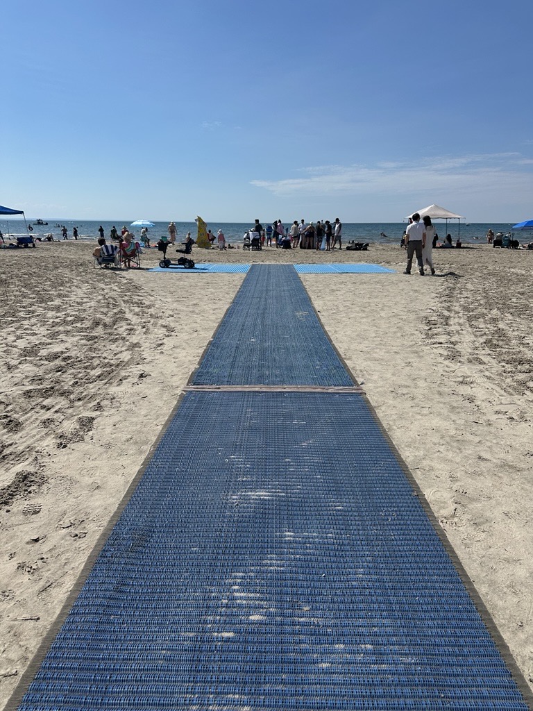 Blue plastic walkway leading from the foreground to a group of beachgoers at the shoreline on a sunny day and calm sea in the background of a crowded beach.